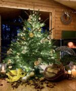 Weihnachtsbaum mit goldenen und silbernen Kugeln, Lichterkette. Vor Kaminfeuer. Im Vordergrund Kaminscheite, goldener Engel und Kerzen. Draußen ist es dunkel,