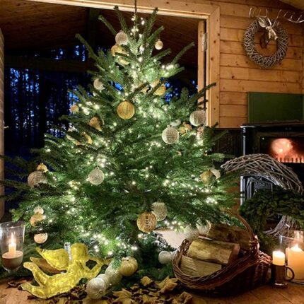 Weihnachtsbaum mit goldenen und silbernen Kugeln, Lichterkette. Vor Kaminfeuer. Im Vordergrund Kaminscheite, goldener Engel und Kerzen. Draußen ist es dunkel,