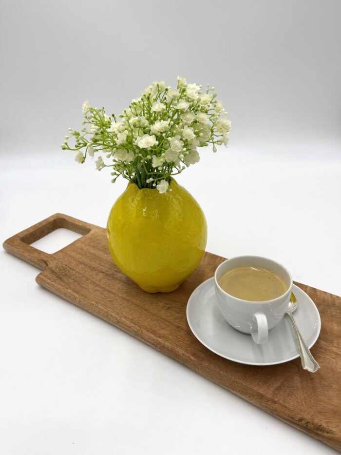 weiße Tasse mit Kaffee, auf Untertasse, gelbe Zitronenvase mit künstlichem Schleierkraut auf Vesperbrett Mangoholz